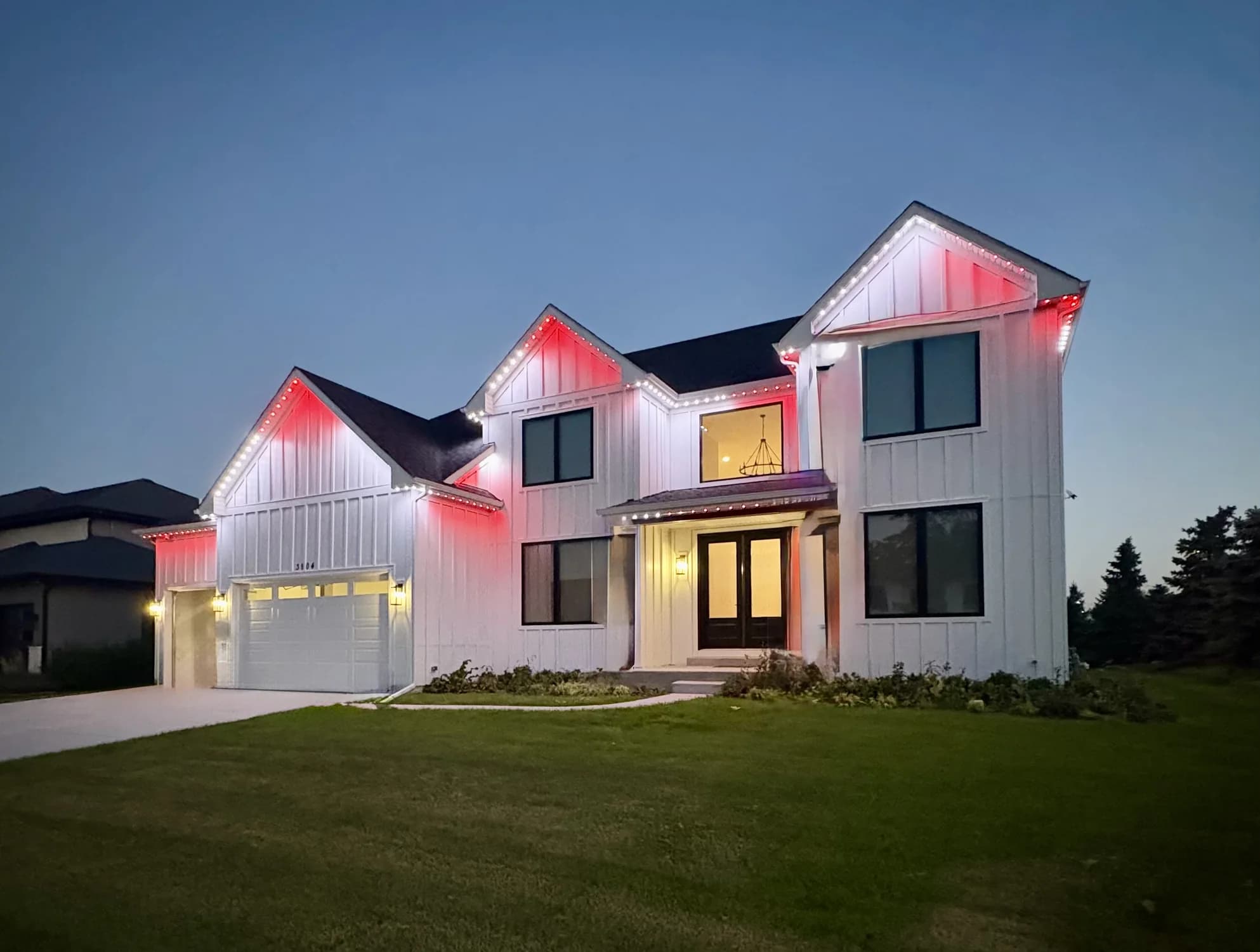 Red accent lighting beneath the peaks of a white modern farmhouse at dusk.